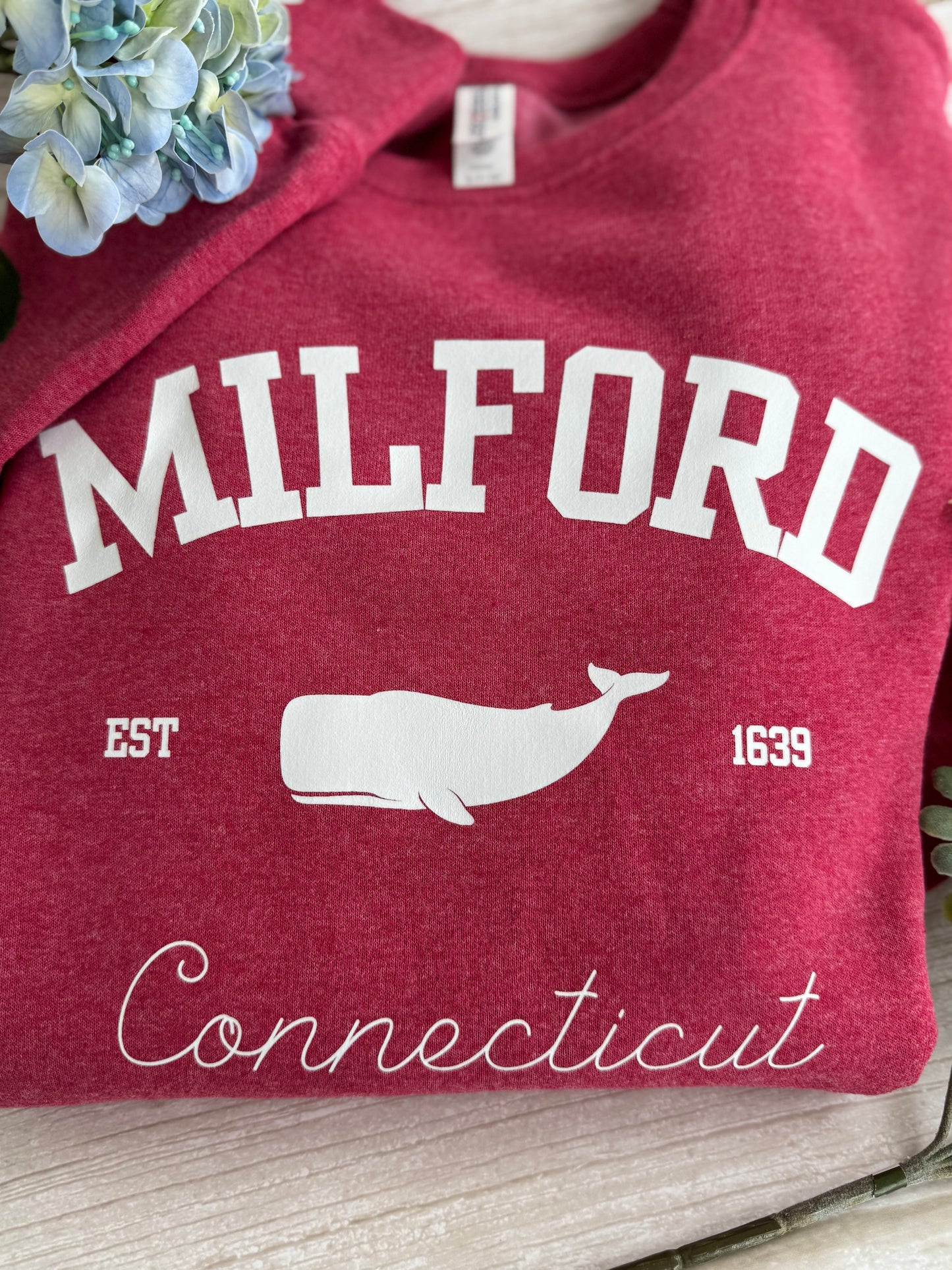 Red sweatshirt with 'Milford, Connecticut' and a whale design on a wooden surface.