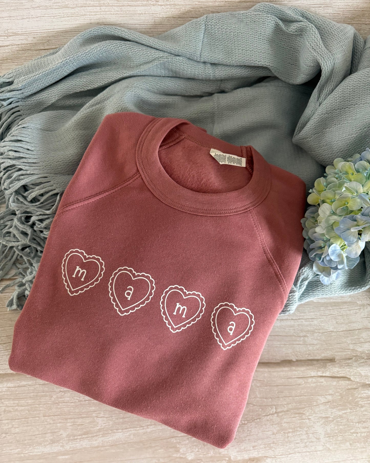 Folded pink sweatshirt with heart designs on a wooden surface with a blue scarf and flowers.