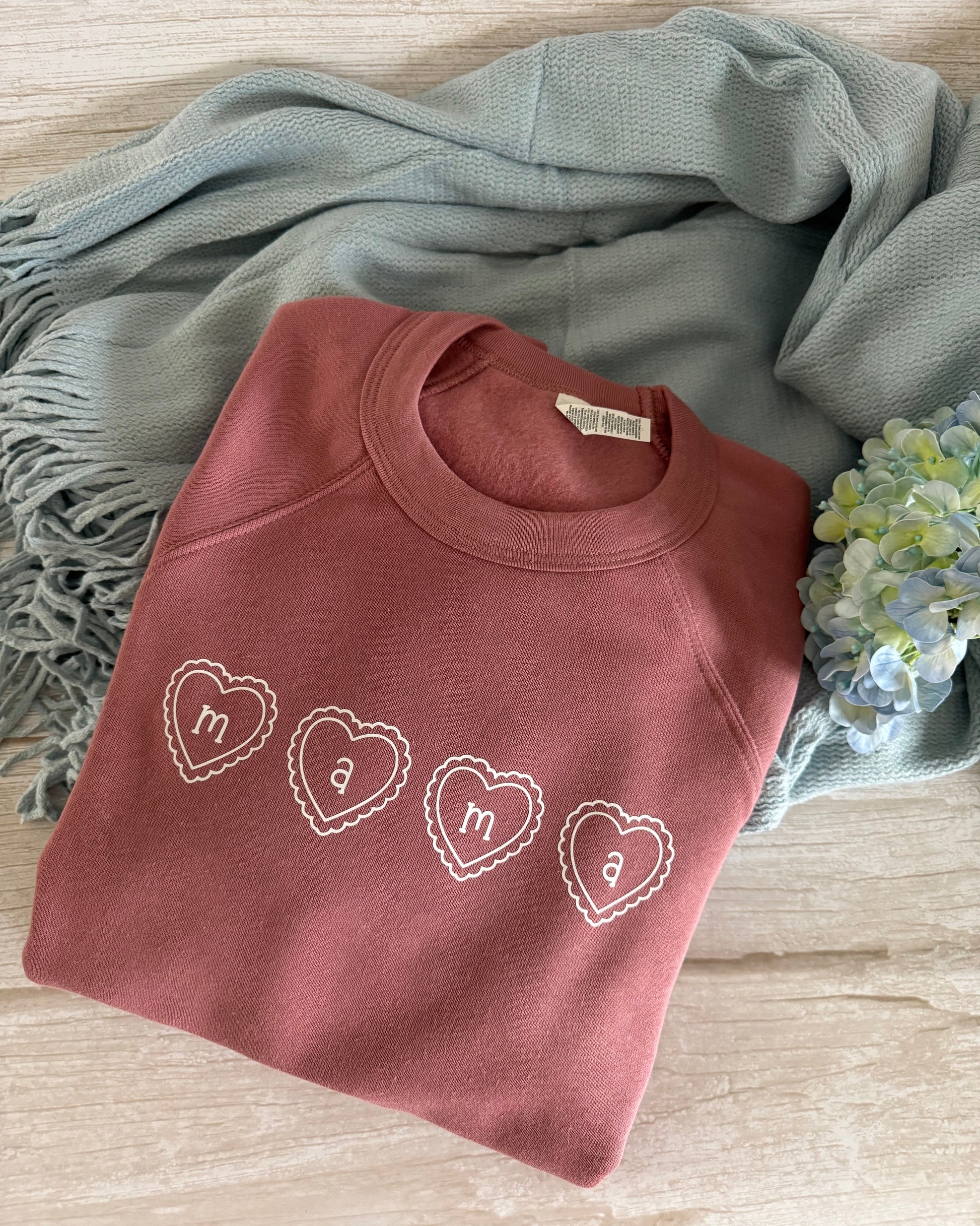 Folded pink sweatshirt with heart designs on a wooden surface with a blue scarf and flowers.