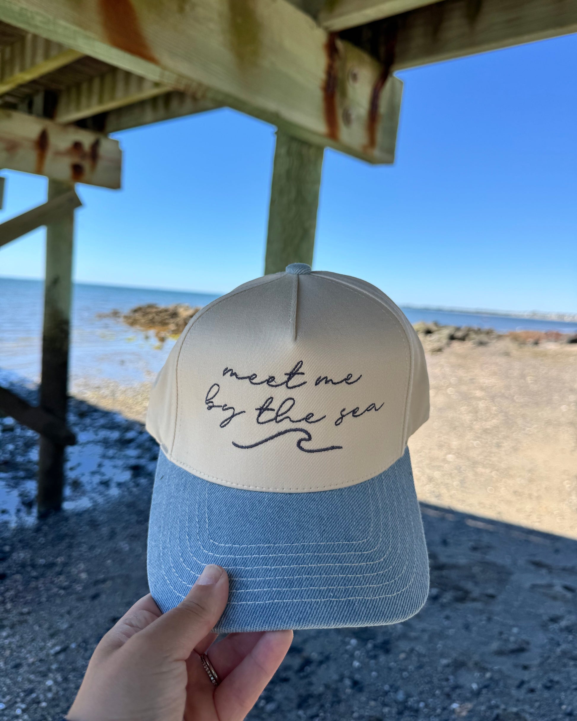 Blue cap with 'meet me by the sea' text held up against a beach backdrop