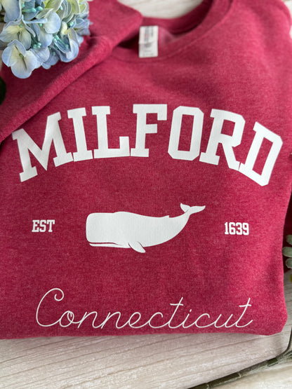 Red sweatshirt with 'Milford, Connecticut' and a whale design on a wooden surface.