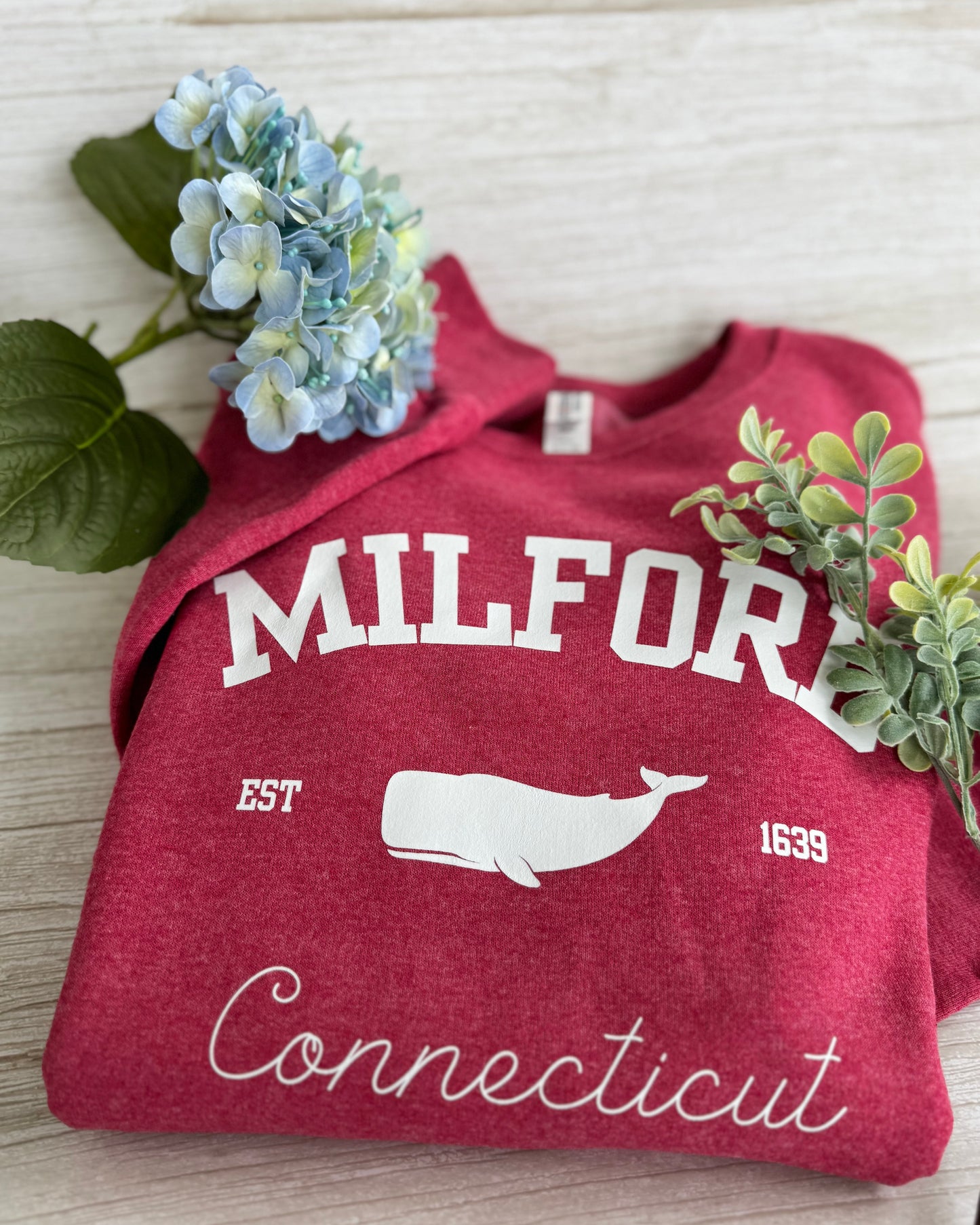 Red sweatshirt with 'Milford, Connecticut' text and whale graphic on a light wooden surface with flowers.