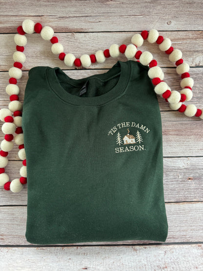forest green crewneck unisex sweatshirt embroidered with a cozy cabin in the woods and the phrase 'tis the damn season for swifties