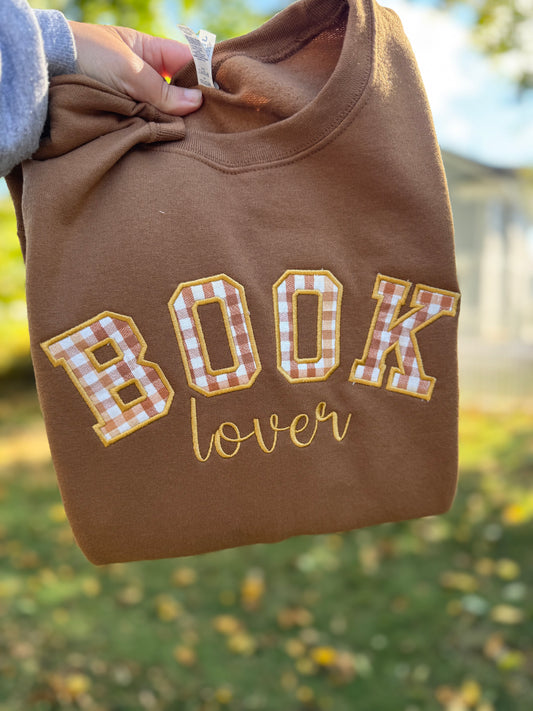The product in the image is a brown sweatshirt. It features the words "BOOK lover" on the front, with "BOOK" written in large, bold letters in a yellow and white checkered pattern and outlined in yellow, while "lover" is written in a smaller, cursive yellow font underneath. The sweatshirt appears to be held up outdoors, with autumn leaves visible in the background.